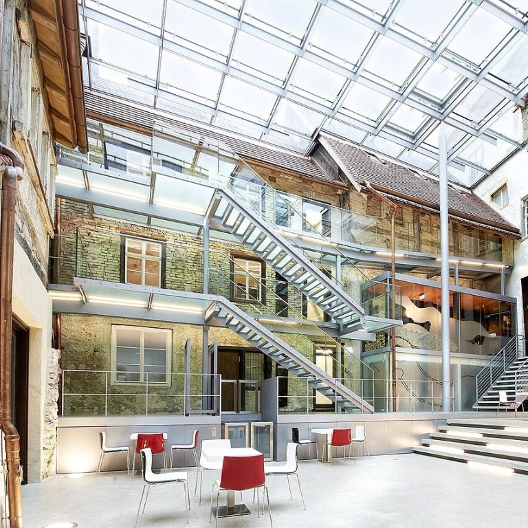 Modernes Atrium zwischen mittelalterlichen Gebäuden mit Glasdach, Treppe und Designer-Möbeln..